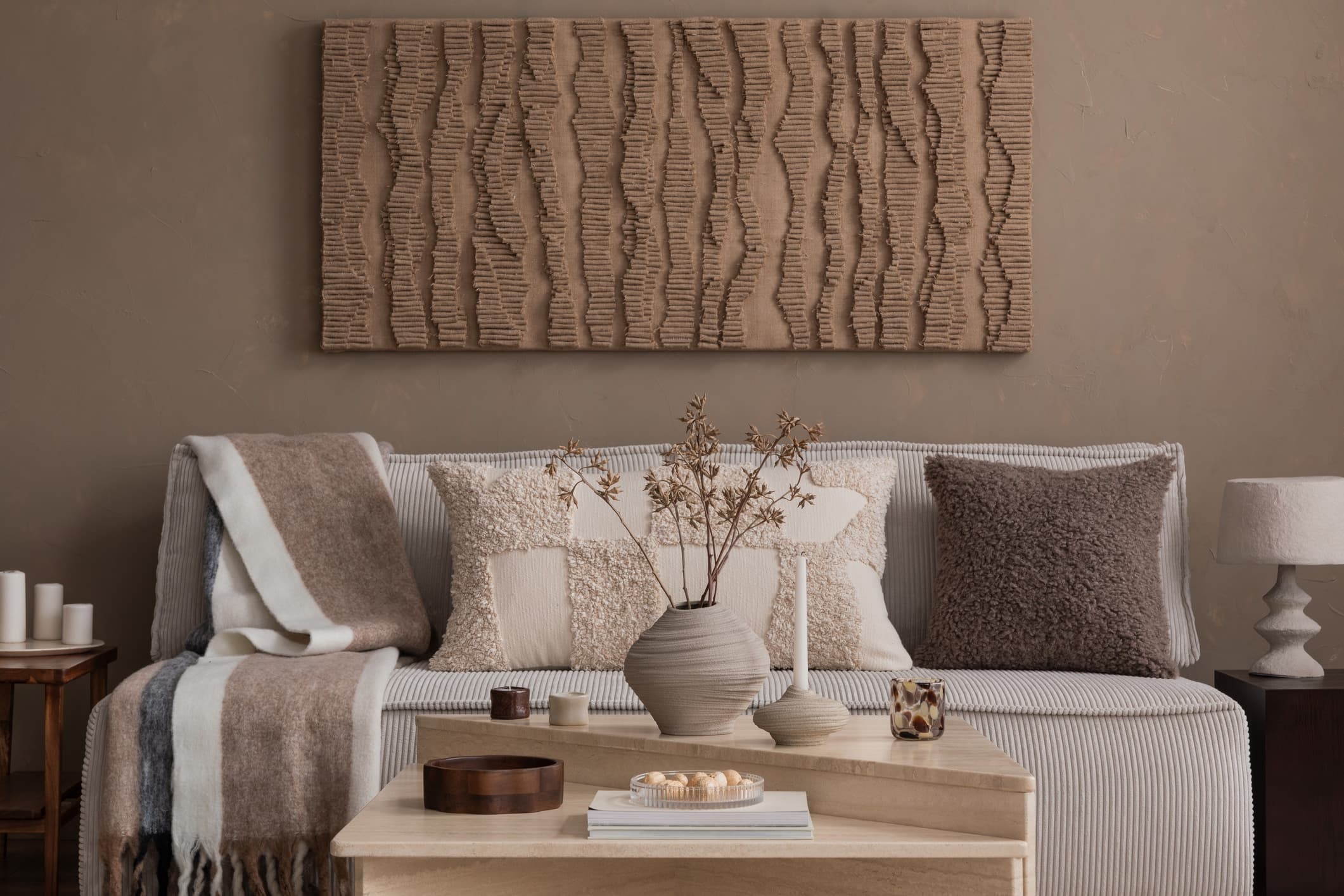 Bildet viser et stilfullt interiør med en sofa i lyse, nøytrale farger med teksturerte puter, et trebord med dekorative gjenstander, og et kunstnerisk veggpanel med bølgende tekstur.