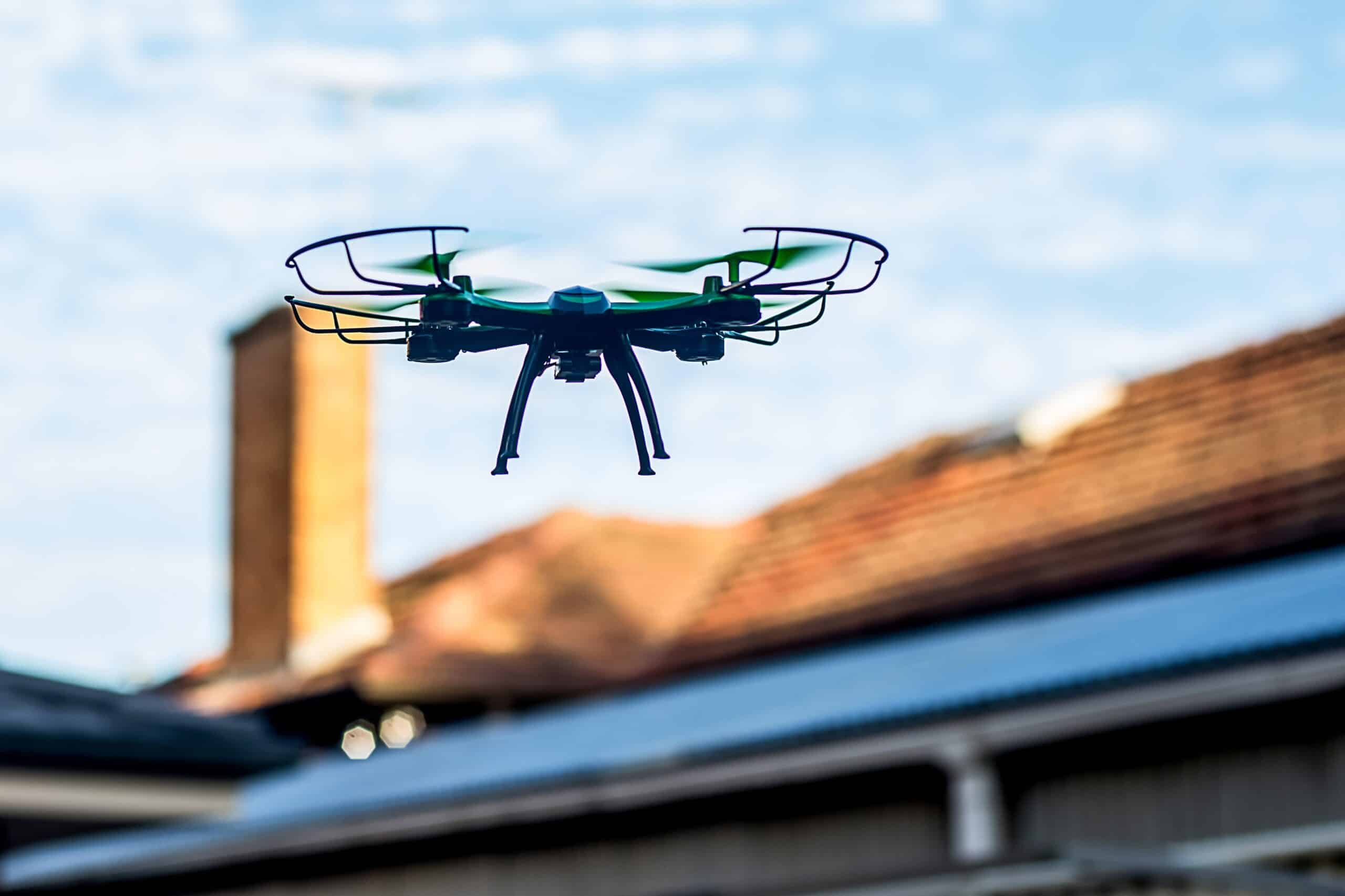 Svart drone svever over røde takstein på et norsk boligområde, klar for å ta dronebilder.